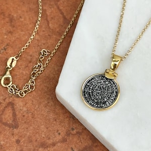 May include: Gold-toned necklace featuring a circular pendant with an intricate, engraved design in black and silver. The pendant is suspended from a delicate gold chain. The necklace rests on a white surface, with a portion of the chain visible.