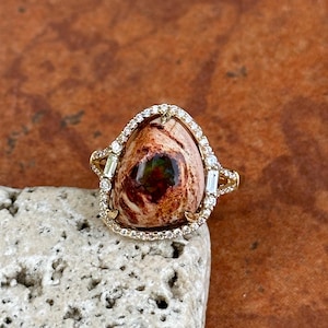 Könnte beinhalten: Ein goldener Ring mit einem großen, ovalen Edelstein mit braunen, roten und grünen Mustern. Der Edelstein ist von kleinen, klaren Diamanten und Baguette-Steinen umgeben, die einen Halo-Effekt erzeugen.