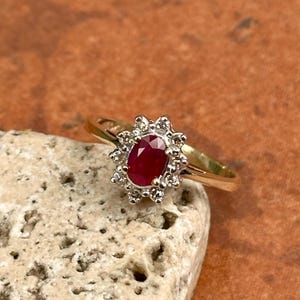 Peut inclure: Une bague en or ornée d'une pierre rouge ovale, entourée d'un halo de petites pierres claires. La bague est mise en valeur sur un fond neutre, soulignant le contraste entre l'or et les pierres scintillantes.
