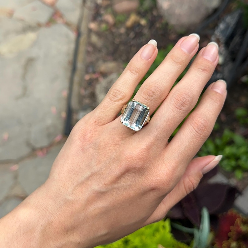 May include: A large, rectangular aquamarine gemstone ring. The light blue gemstone is set in a gold band with intricate detailing. The ring is displayed on a hand with manicured nails, showcasing the ring's size and design.