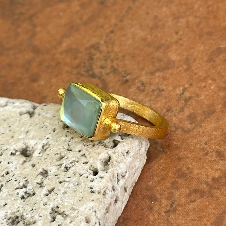 May include: A gold-toned ring with a square, pale green gemstone. The ring has a textured band and small, decorative spheres on either side of the gemstone setting. The ring is displayed on a textured, off-white surface.
