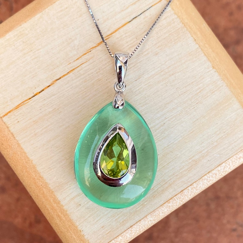 May include: A silver pendant necklace featuring a pear-shaped, light green gemstone with a smaller, olive-green gemstone set inside. The pendant hangs from a delicate silver chain. The necklace is displayed on a wooden surface.
