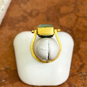 May include: A gold-toned ring with a rectangular, light-green gemstone. The ring features a unique design with a curved, open setting that cradles a white, rounded form. The ring is displayed on a white surface against a warm, brown background.