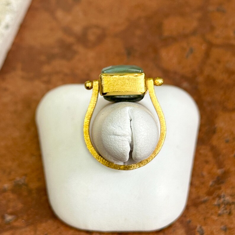 May include: A gold-toned ring with a rectangular, light-green gemstone. The ring features a unique design with a curved, open setting that cradles a white, rounded form. The ring is displayed on a white surface against a warm, brown background.