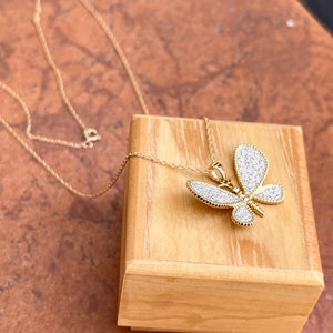 May include: A gold-colored butterfly pendant necklace with a delicate chain. The butterfly is encrusted with small, sparkling white stones. The necklace is displayed on a small wooden box, set against a warm, textured background.