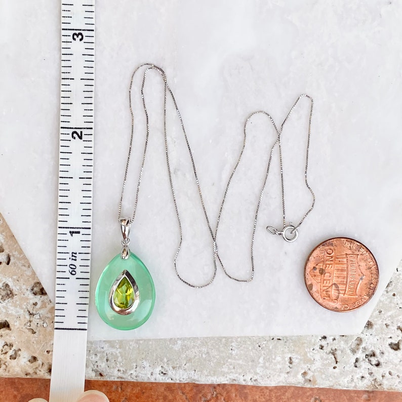 May include: A silver necklace with a pendant featuring a light green teardrop-shaped stone with a smaller, yellow-green stone inside. The necklace is next to a ruler showing measurements in inches and a penny.