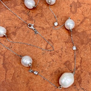 K&ouml;nnte beinhalten: Eine elegante Halskette mit gro&szlig;en, wei&szlig;en Perlen und kleinen, funkelnden, klaren Kristallen an einer feinen Silberkette. Die Perlen sind entlang der Kette angeordnet und erzeugen einen raffinierten Look. Die Halskette wird vor einem warmen, strukturierten Hintergrund gezeigt.