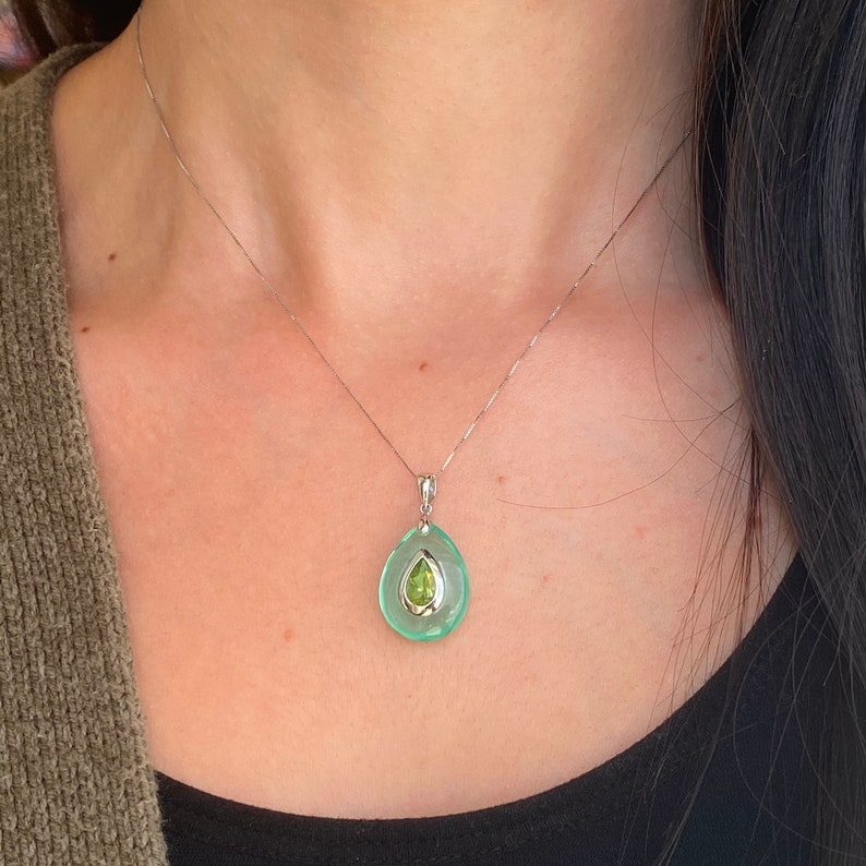 May include: A silver necklace with a pendant featuring a teardrop-shaped, light green stone with a smaller, pear-shaped, light green stone in the center. The necklace is worn around the neck.