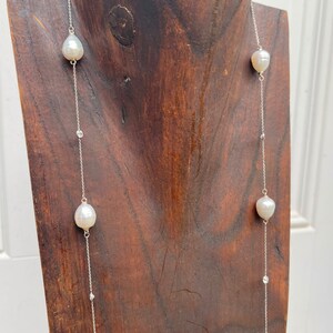 K&ouml;nnte beinhalten: Eine lange, zarte Halskette mit einer silbernen Kette und gro&szlig;en, wei&szlig;en, tropfenf&ouml;rmigen Perlen. Kleine, klare Kristalle sind entlang der Kette eingestreut. Die Halskette wird auf einem dunkelbraunen Holzst&auml;nder pr&auml;sentiert.