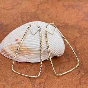 May include: A pair of gold triangle-shaped hoop earrings with a textured finish. The earrings are displayed on a white seashell.
