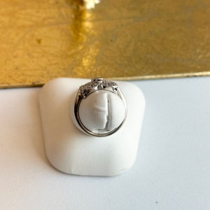 May include: A silver ring with a clear stone and a diamond-like design. The ring is on a white display stand.