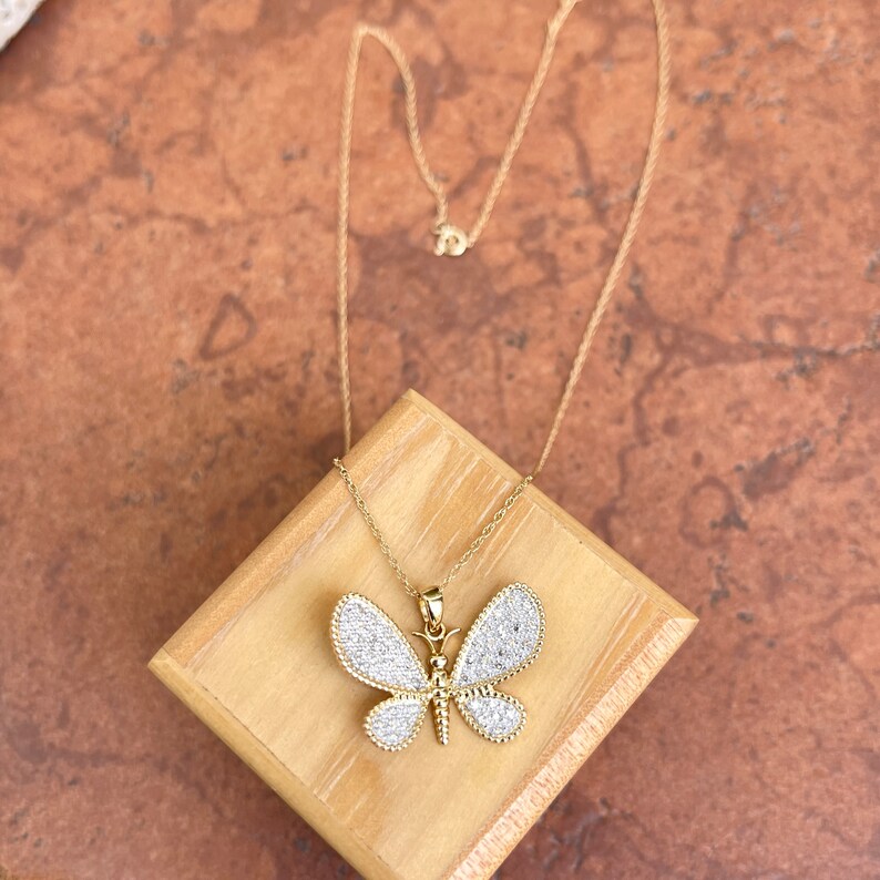 May include: A gold-colored butterfly pendant necklace with a delicate chain. The butterfly pendant is adorned with small, sparkling white stones, creating a dazzling effect. The necklace is displayed on a wooden block against a textured, brown surface.