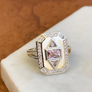 May include: Silver-toned ring with a rectangular face. The face features a central rectangular pink gemstone, surrounded by smaller pink stones and a geometric design. The ring is set on a white surface.