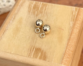 May include: Two small, gold-colored, round earrings with a loop at the bottom, resting on a light brown wooden surface. The earrings have a shiny, polished finish. The background is a light brown wooden box.