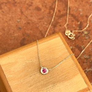 May include: A delicate gold necklace featuring a small, round, pink gemstone pendant. The pendant is set in a bezel setting and hangs from a fine gold chain. The necklace is displayed on a wooden surface.