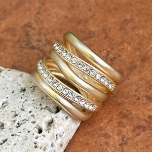 May include: A gold-toned ring with multiple bands, some adorned with small, clear crystals. The ring has a layered design, creating a textured and eye-catching appearance. The ring is set against a neutral background.