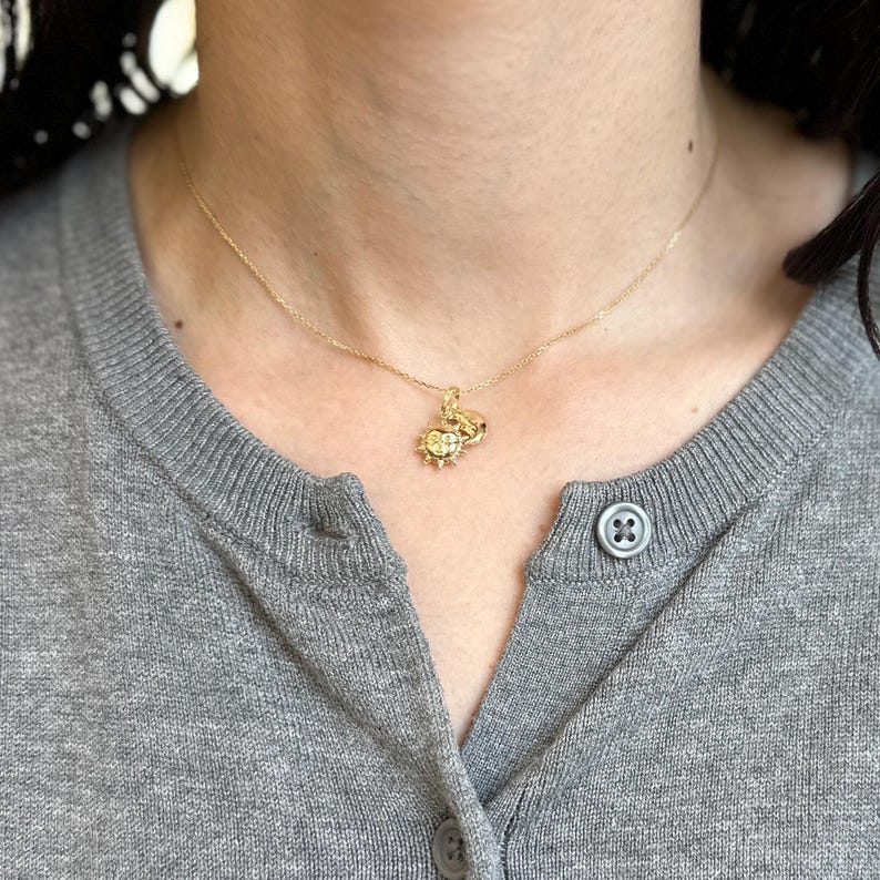 May include: A gold-colored necklace with a pendant featuring a sun design. The necklace is worn over a gray cardigan. The pendant has a detailed sunburst design with a central element.
