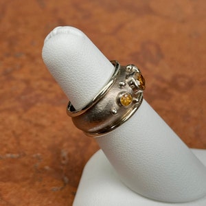 May include: A silver ring with three round, yellow gemstones and small silver accents. The ring is displayed on a white finger form against a warm, neutral background. The ring's design features a textured band and a polished outer edge.