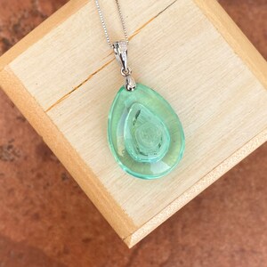 May include: A silver necklace with a pendant featuring a layered, teardrop-shaped, light green glass design. The pendant has a textured, swirling pattern in the center. The necklace is displayed on a wooden surface.
