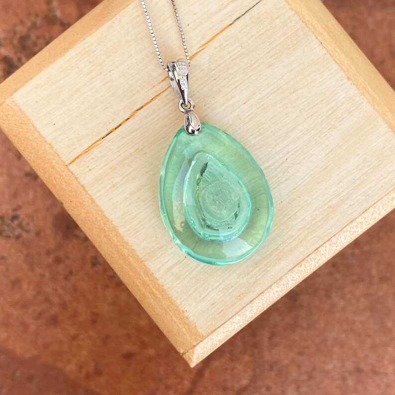 May include: A silver necklace with a pendant featuring a layered, teardrop-shaped, light green glass design. The pendant has a textured, swirling pattern in the center. The necklace is displayed on a wooden surface.