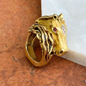 Könnte beinhalten: Ein goldener Ring in Form eines Pferdekopfes mit texturierter Mähne. Das Pferd hat einen kleinen roten Edelstein als Auge und einen einzelnen klaren, diamantähnlichen Stein auf der Stirn. Das Metall hat ein gebürstetes Finish mit dunklen Akzenten.