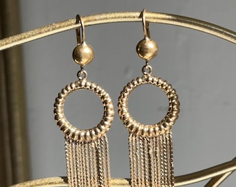 May include: A pair of gold-colored earrings. Each earring features a small, round stud, a textured circular hoop, and a fringe of delicate chains. The earrings are displayed against a gold-colored, curved metal structure.