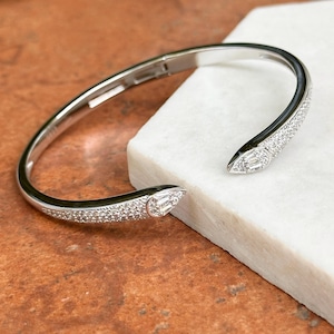 Pode incluir: Uma pulseira de punho aberta prateada. A pulseira apresenta uma fileira de pequenas pedras transparentes ao longo das bordas externas. As extremidades da pulseira têm um design pontiagudo com uma pedra retangular clara maior. A pulseira é exibida em uma superfície branca.