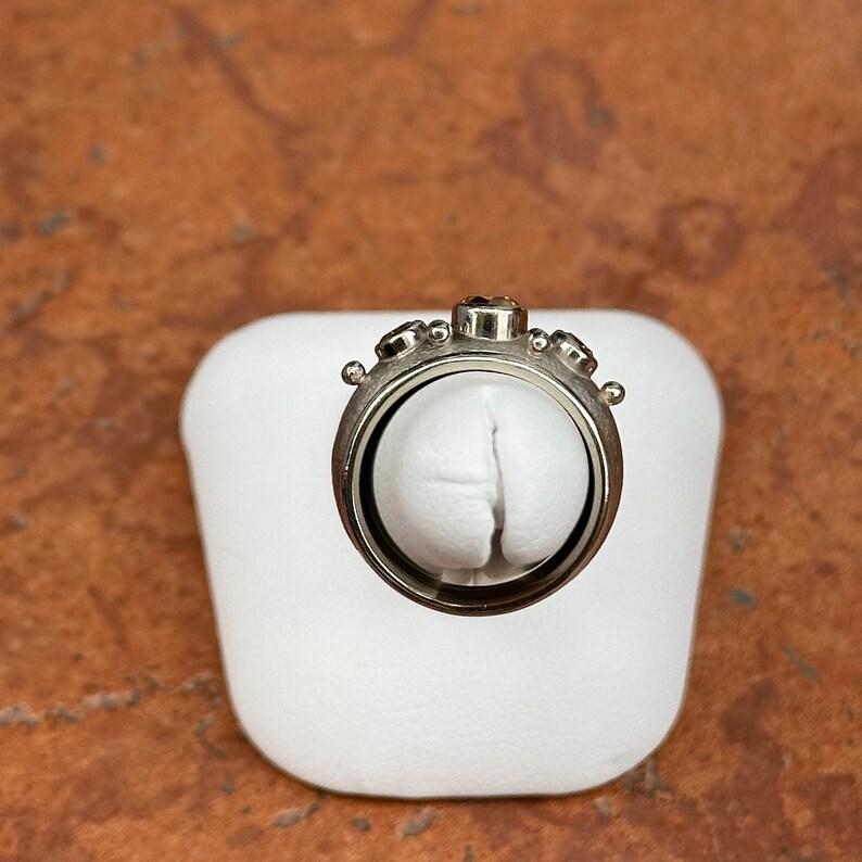 May include: A silver-toned ring with a unique design, featuring a central dark gemstone and smaller stones on the sides. The ring is displayed on a white surface against a warm, textured background.