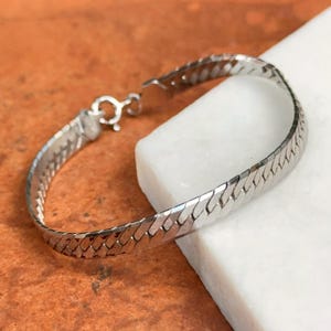 May include: A silver-colored herringbone bracelet with a lobster clasp. The bracelet is displayed on a white surface against a brown background. The bracelet's design features a series of interconnected, flat, rectangular links.