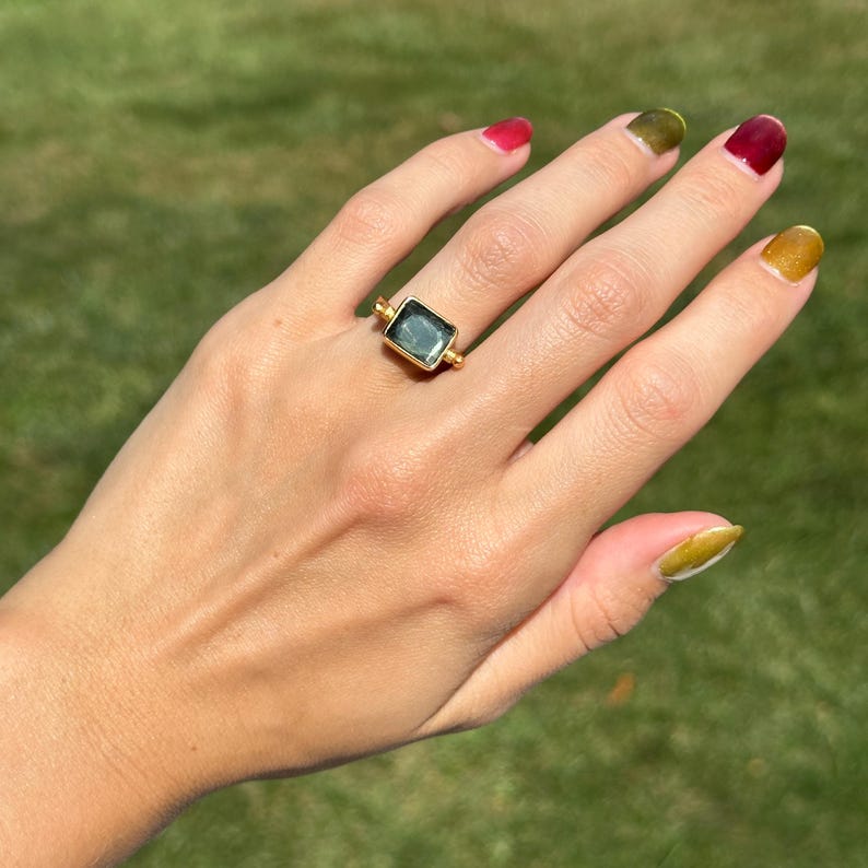 May include: A gold ring with a rectangular, teal-colored gemstone. The ring is set on a hand with painted nails in shades of red, green, and gold. The background is a blurred green.