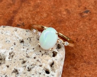May include: A gold ring featuring an oval, light-colored opal gemstone. The ring has small, clear accent stones on either side of the opal. The ring is set on a light-colored, textured surface.