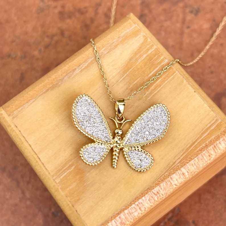 May include: A gold-toned butterfly pendant necklace with a delicate chain. The butterfly wings are encrusted with small, sparkling stones, and the edges are outlined with a beaded design. The necklace rests on a wooden box.
