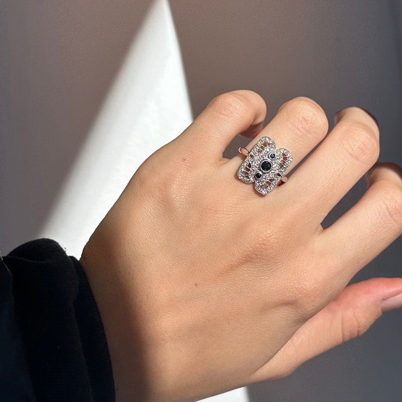 May include: A silver ring with a delicate, intricate design featuring a central cluster of black gemstones surrounded by smaller clear stones.