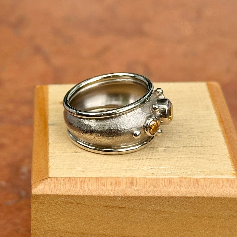 May include: A silver ring with a textured band and two small, round, yellow gemstones. The ring has a wide band with a textured surface and a polished edge. The gemstones are set on the side of the ring.