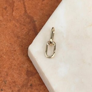 May include: A small, gold-colored jewelry clasp with an oval-shaped loop and a hinged top. The clasp is displayed on a white surface with a brown background. The clasp is likely used for necklaces or bracelets.