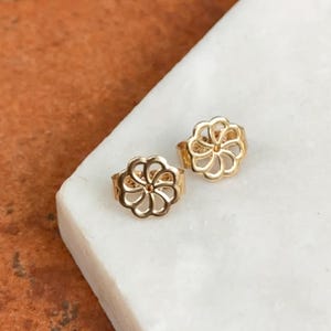 May include: A pair of gold-toned earring backs featuring a circular, open-work floral design with eight rounded petals radiating from a central point. These metal earring backs are designed to secure stud earrings and have a polished finish.