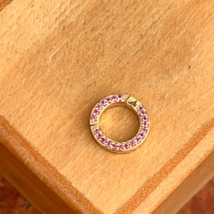 May include: A gold-coloured, circular clasp with a ring of small, pink gemstones. The clasp is set against a light brown wooden surface. The clasp is designed to secure jewellery. The gemstones are a vibrant pink.
