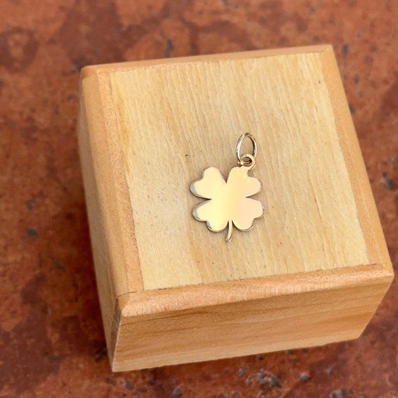Puede incluir: Un colgante de tr&eacute;bol de cuatro hojas de oro. El colgante est&aacute; sobre una superficie de madera.