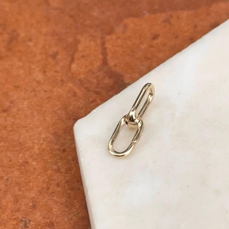 May include: A small, gold-colored jewelry link with an oval and a rectangular shape. The link is shown on a white marble surface with a brown background.