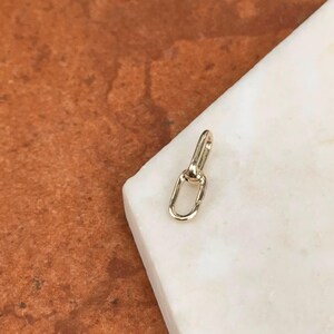 May include: A small, gold-colored jewelry clasp with an oval-shaped loop and a connecting bar. The clasp is set against a white marble surface and a brown background.