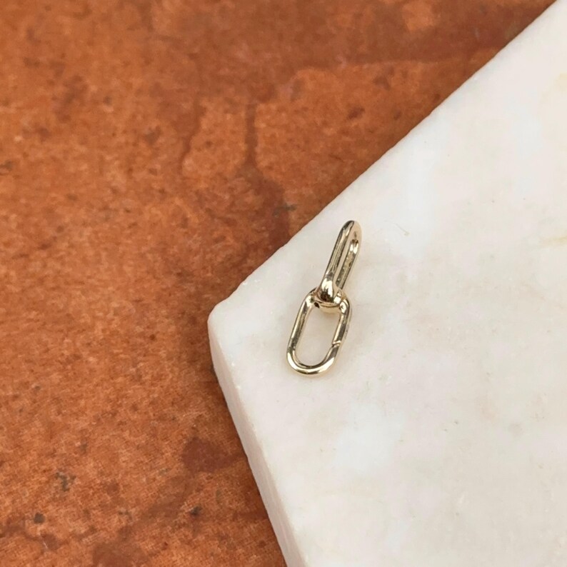 May include: A small, gold-colored jewelry clasp with an oval-shaped loop and a connecting bar. The clasp is set against a white marble surface and a brown background.