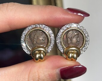 May include: A pair of gold and silver earrings with a coin design. Each earring features a round coin with a profile of a woman's face, surrounded by a circle of small diamonds. The coins are attached to a gold loop with a knot design.