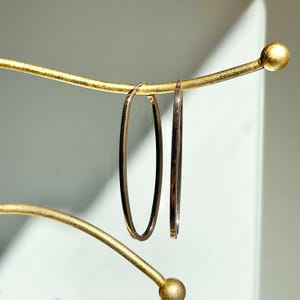 Op de afbeelding: Twee goudkleurige ovale oorringen hangen aan een goudkleurige displaystandaard. De oorbellen zijn dun en hebben een gepolijste afwerking. De standaard heeft een gebogen ontwerp met een bal aan het uiteinde.