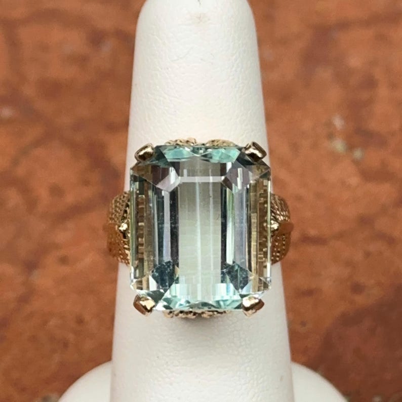 May include: A gold ring featuring a large, rectangular, light blue gemstone. The gemstone is set in a detailed gold band with ornate filigree detailing on the sides. The ring is displayed on a white finger.