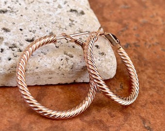 May include: A pair of rose gold hoop earrings with a twisted rope design. The earrings are displayed on a textured, off-white stone against a brown and orange surface. The earrings are a classic jewelry accessory.