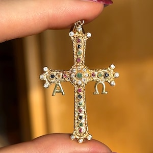 Può includere: Un ciondolo a forma di croce dorata con dettagli intricati. La croce è adornata con varie pietre preziose rosse, verdi, nere e trasparenti. Le lettere "A" e "Ω" sono visibili.