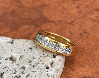 Op de afbeelding: Een gouden ring met een rechthoekig ontwerp, met twee rijen vierkant geslepen, heldere edelstenen. De ring is geplaatst op een lichtgekleurde, getextureerde steen, met een warme, roodbruine achtergrond.