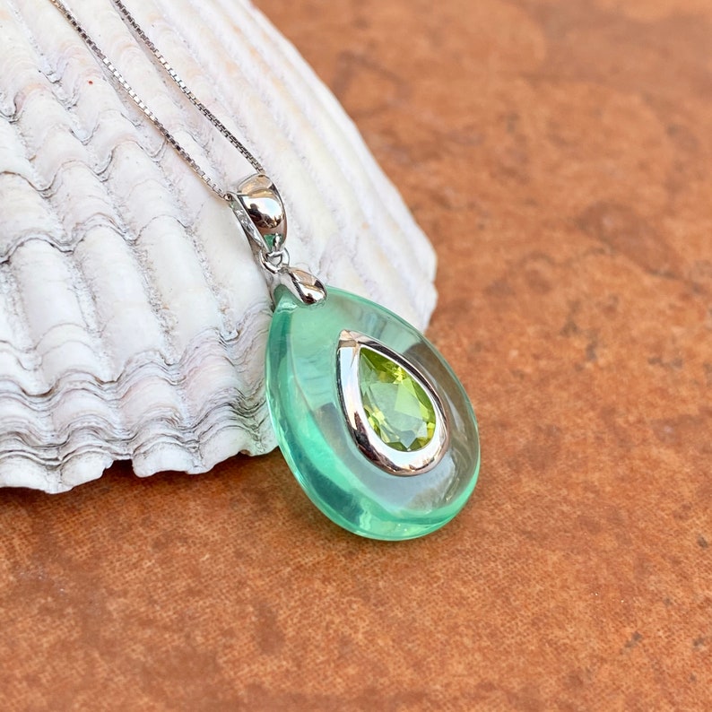 May include: A silver necklace with a pear-shaped pendant. The pendant has a light green outer layer with a silver-colored trim, and a smaller, pear-shaped, light green gemstone in the center. The necklace is displayed on a white seashell.