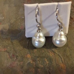 May include: A pair of elegant drop earrings featuring large, lustrous white pearls. The earrings have a silver-toned, curved hook design. The pearls are teardrop-shaped and have a smooth, reflective surface. The earrings are displayed on a white, rectangular backing.