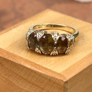 May include: A gold ring featuring three oval-cut gemstones in shades of brown and green, accented with small, sparkling diamonds. The ring is set on a wooden surface, showcasing its elegant design and craftsmanship. A classic piece of jewelry.